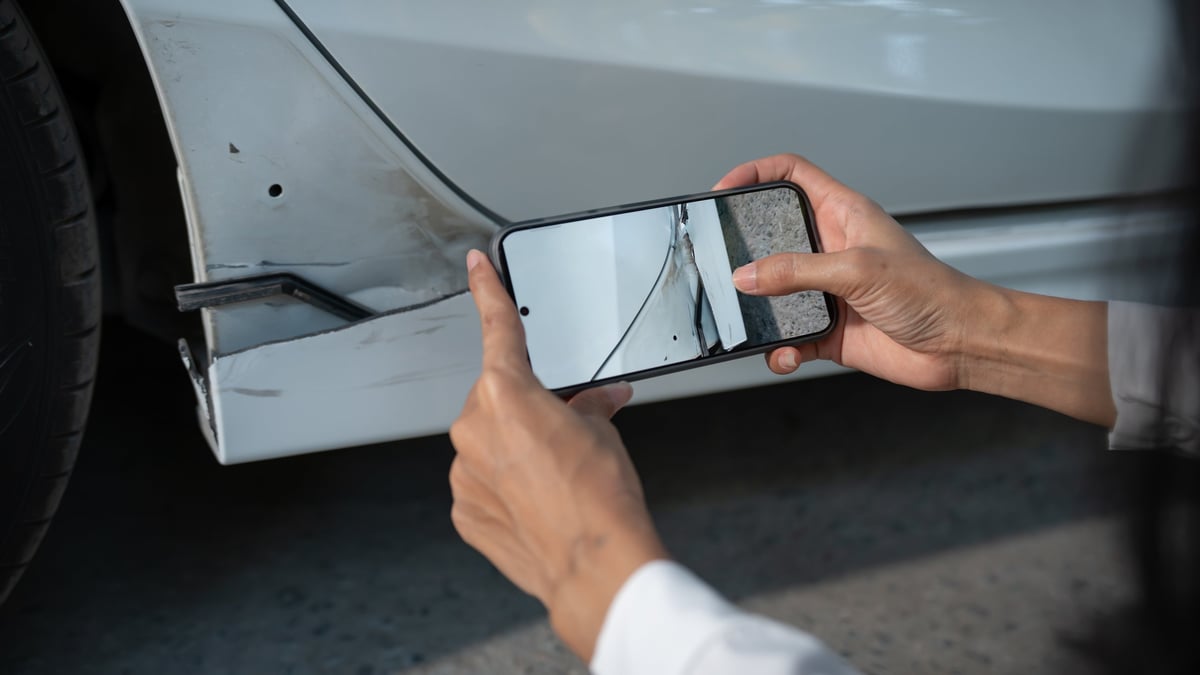 Person using a smartphone to photograph car body damage and a dent on a white vehicle, documenting the condition for an insurance claim or repair estimate
